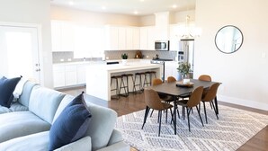 A modern kitchen with white cabinets, a central island with stools, a dining table with chairs, and a hanging light fixture.