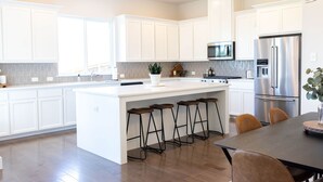 A modern kitchen with white cabinetry, a central island with stools, stainless steel appliances, and a large window.