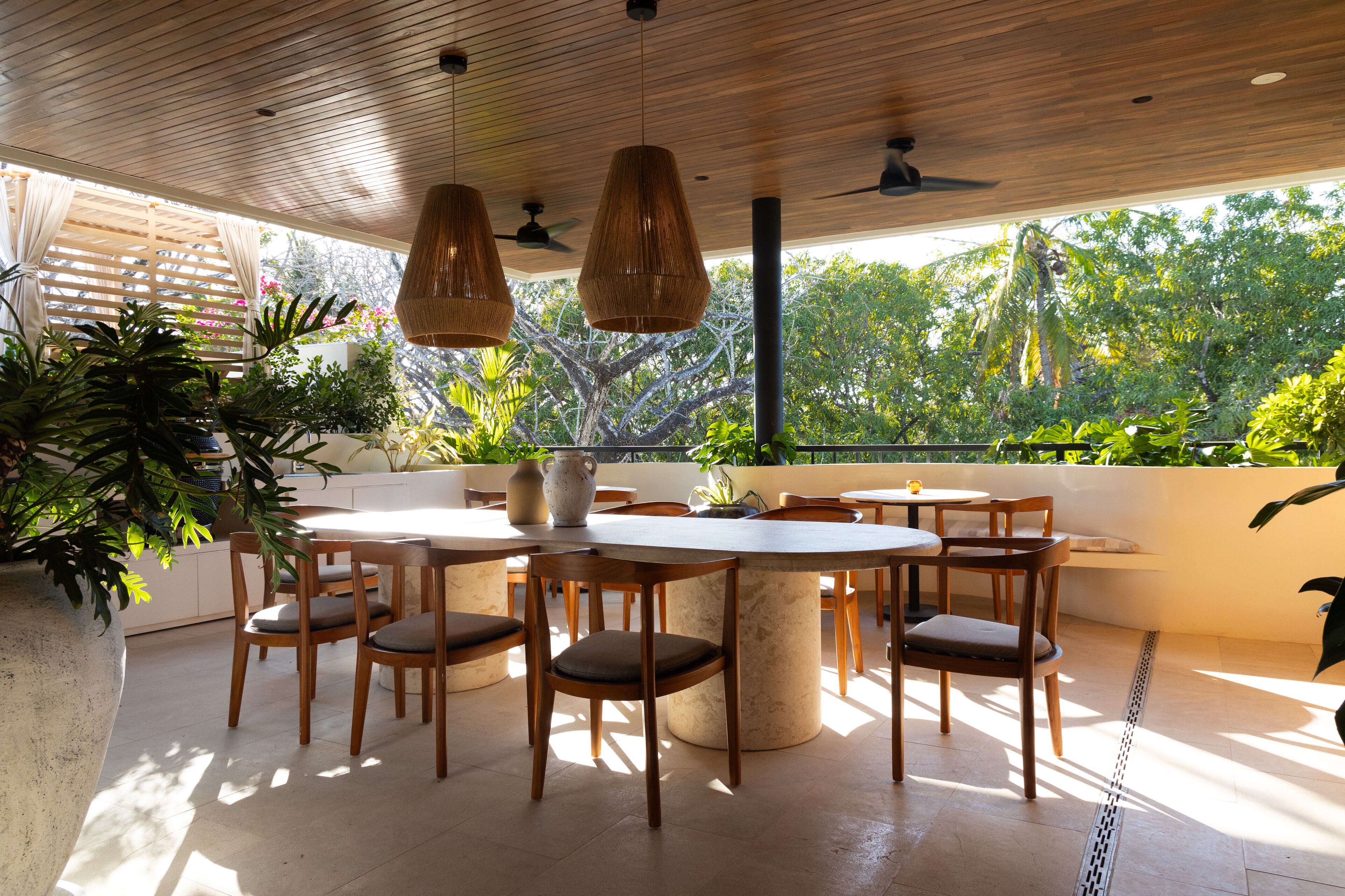 A modern outdoor dining area with wooden furniture and hanging lights, surrounded by lush greenery and large windows.