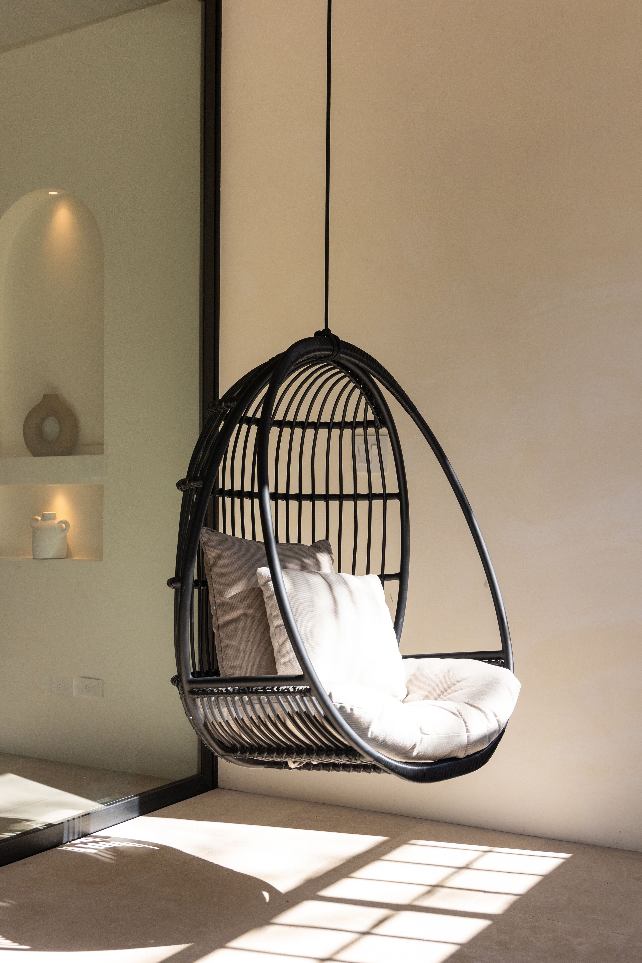 Un fauteuil suspendu avec des coussins, placé dans une pièce dotée d’un grand miroir et d’une étagère ornée d’objets décoratifs.
