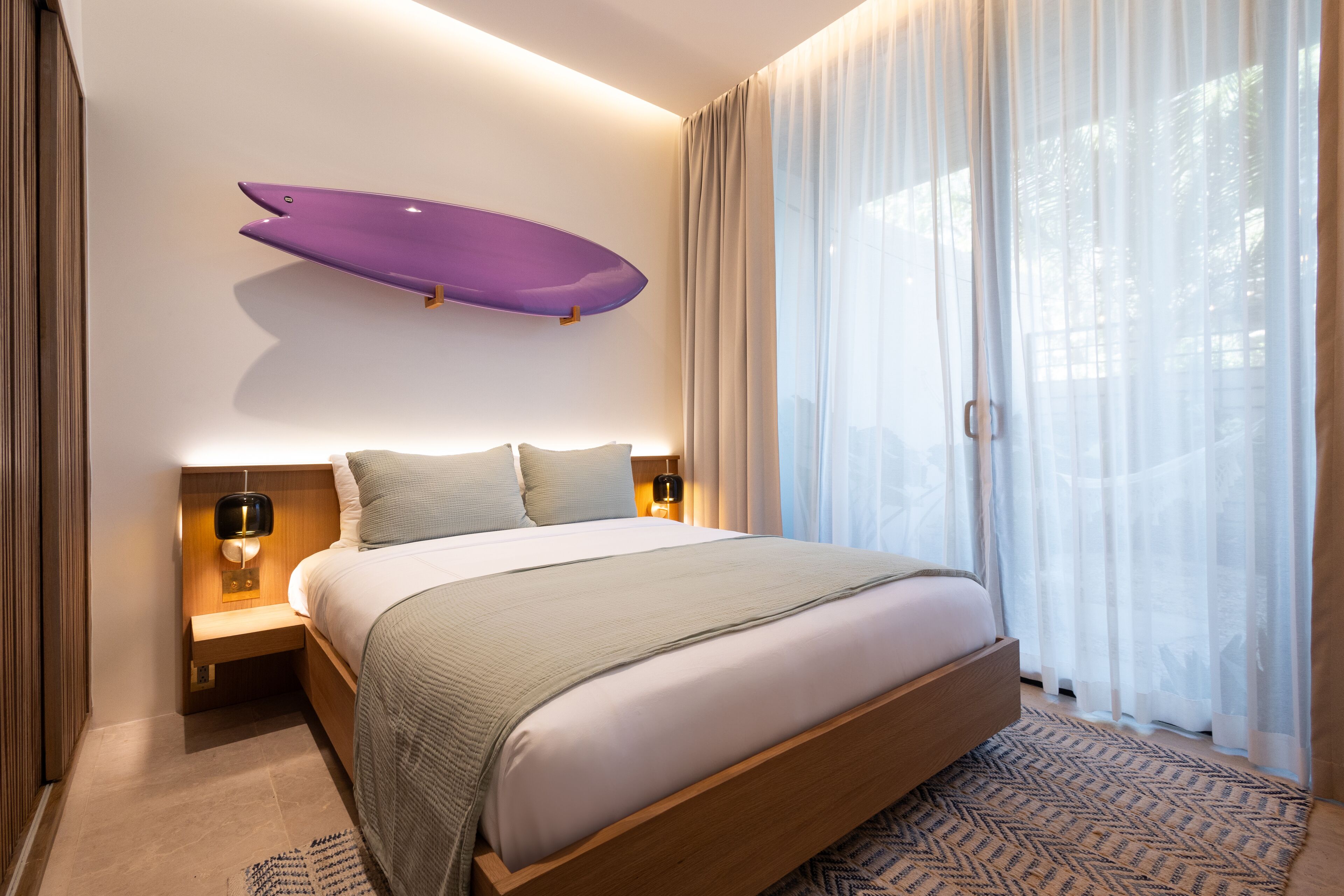 Ett modernt hotellrum med en stor säng, en surfbräda som hänger på väggen och utsikt genom tunna gardiner.