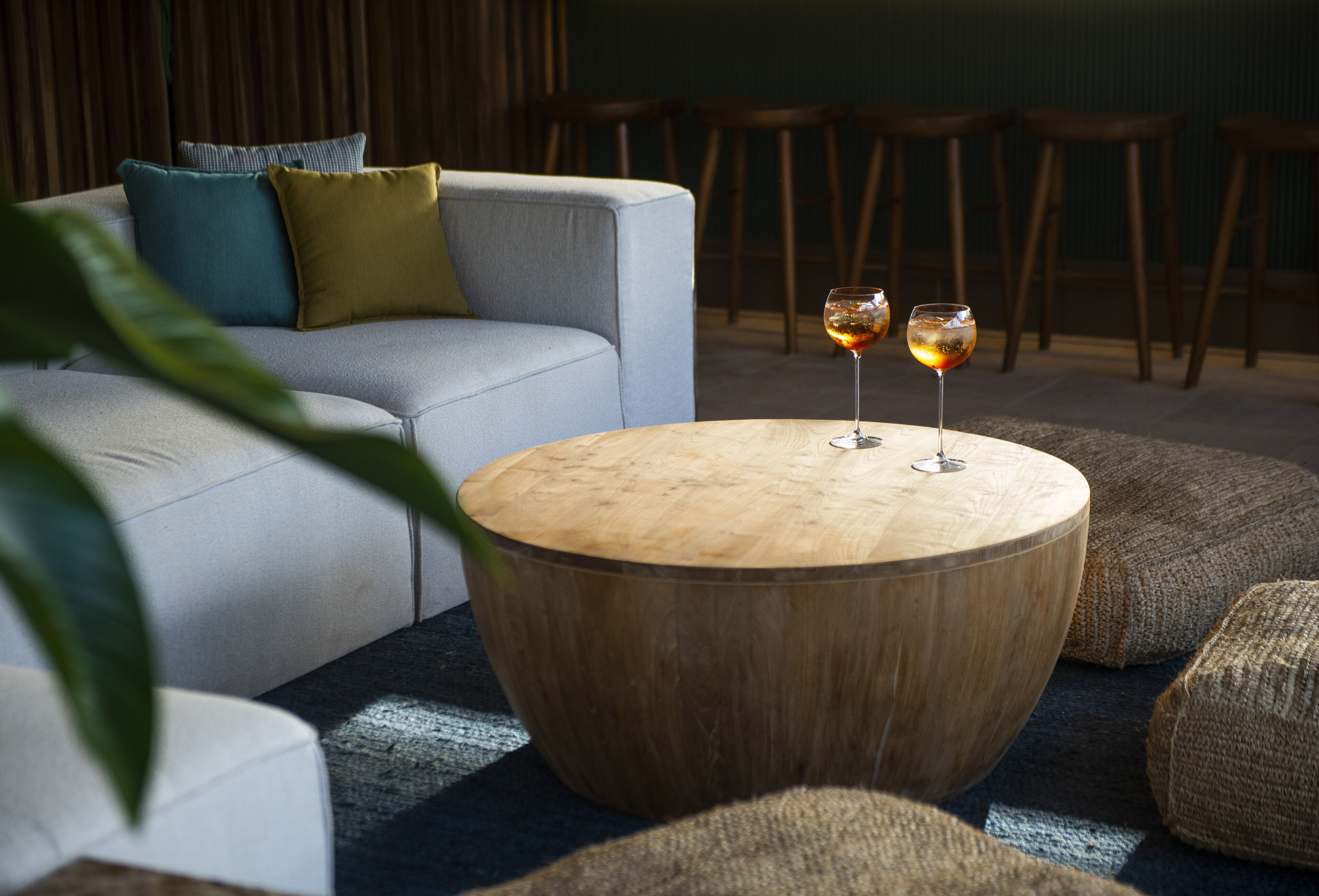 Una mesa redonda de madera con dos copas de vino, un sofá gris con cojines y una planta en primer plano.