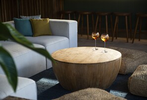 A round wooden coffee table with two glasses of wine, a grey sofa with cushions, and a plant in the foreground.