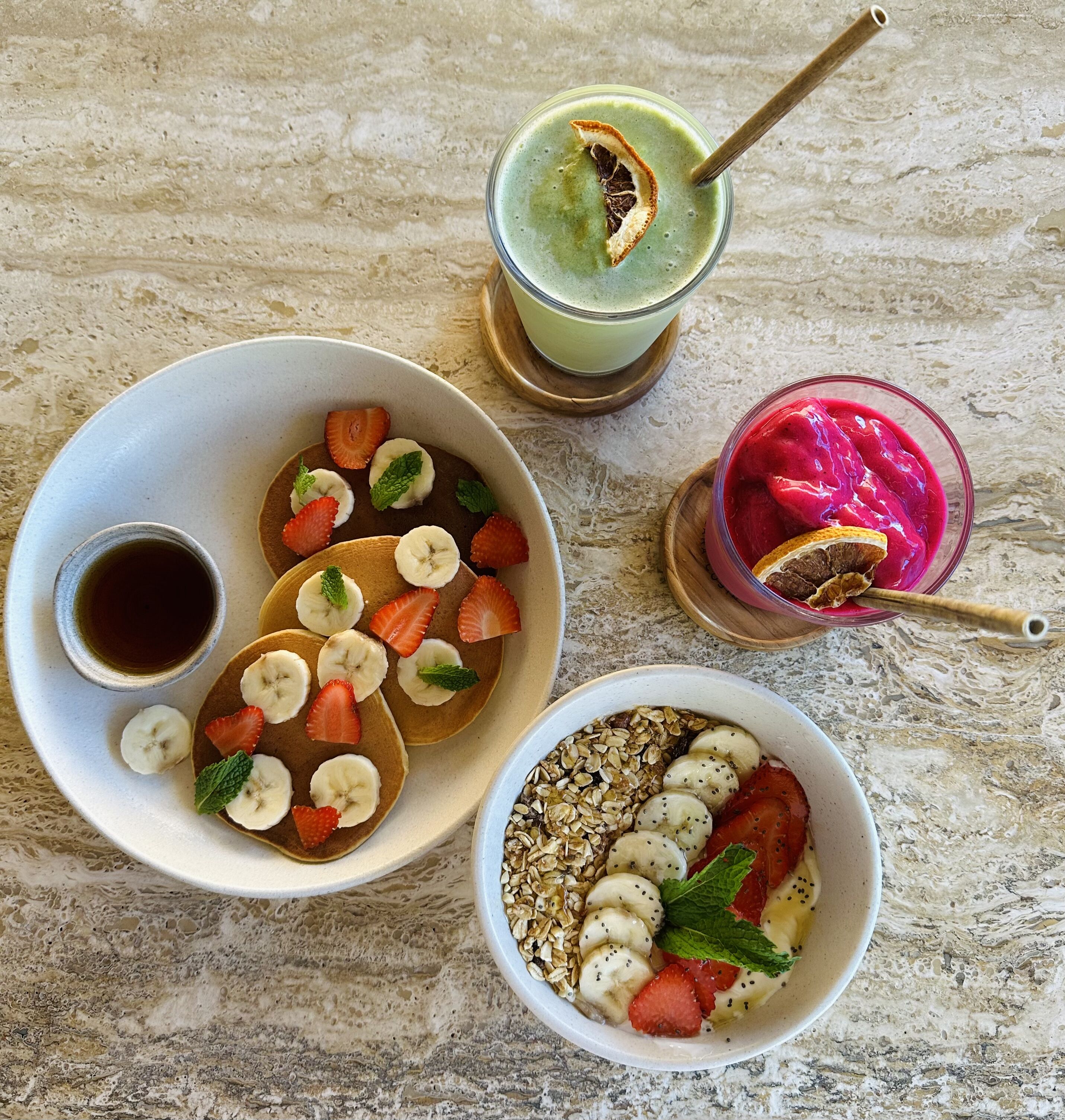 Een ontbijttafel met pannenkoeken, belegd met bananen en aardbeien, een kom granola met bananen en aardbeien, en twee smoothies met fruitdecoratie.