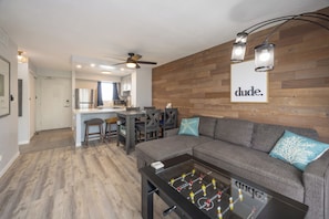 A living room with a foosball table, a sofa with decorative pillows, a wooden wall, and a kitchen area in the background. - Spacious Modern 2Bdrm Condo, Free Parking in Bldg., Full Kitchen, Sleeps 6. (Honolulu)