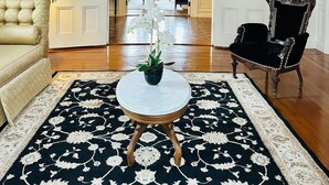 A spacious room with a large chandelier, a round table with a potted orchid, a cream-colored sofa, and a dark upholstered chair. - The Hamilton Hoppin House (Middletown)