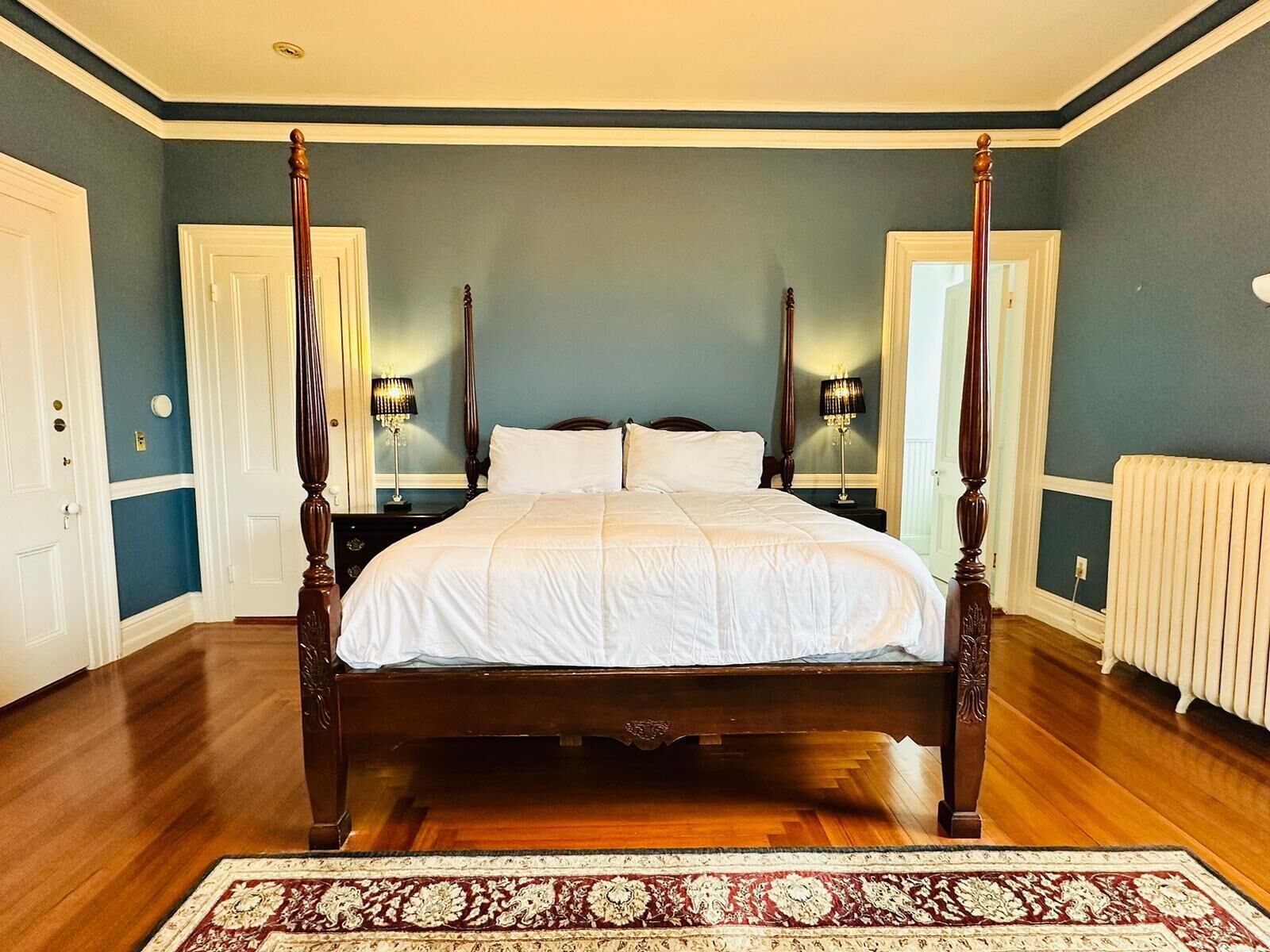 A four-poster bed with white linens, a dark wooden floor, and a patterned rug.