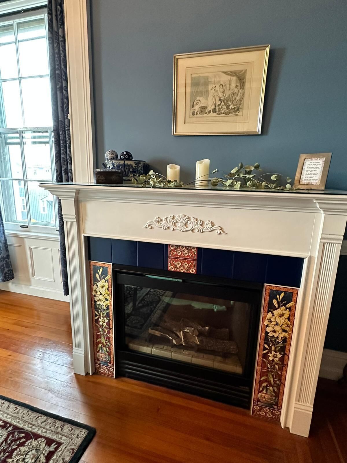 Una chimenea con azulejos decorativos, una repisa con obras de arte enmarcadas y una ventana con cortinas.