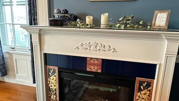 Una chimenea con azulejos decorativos, una repisa con obras de arte enmarcadas y una ventana con cortinas.