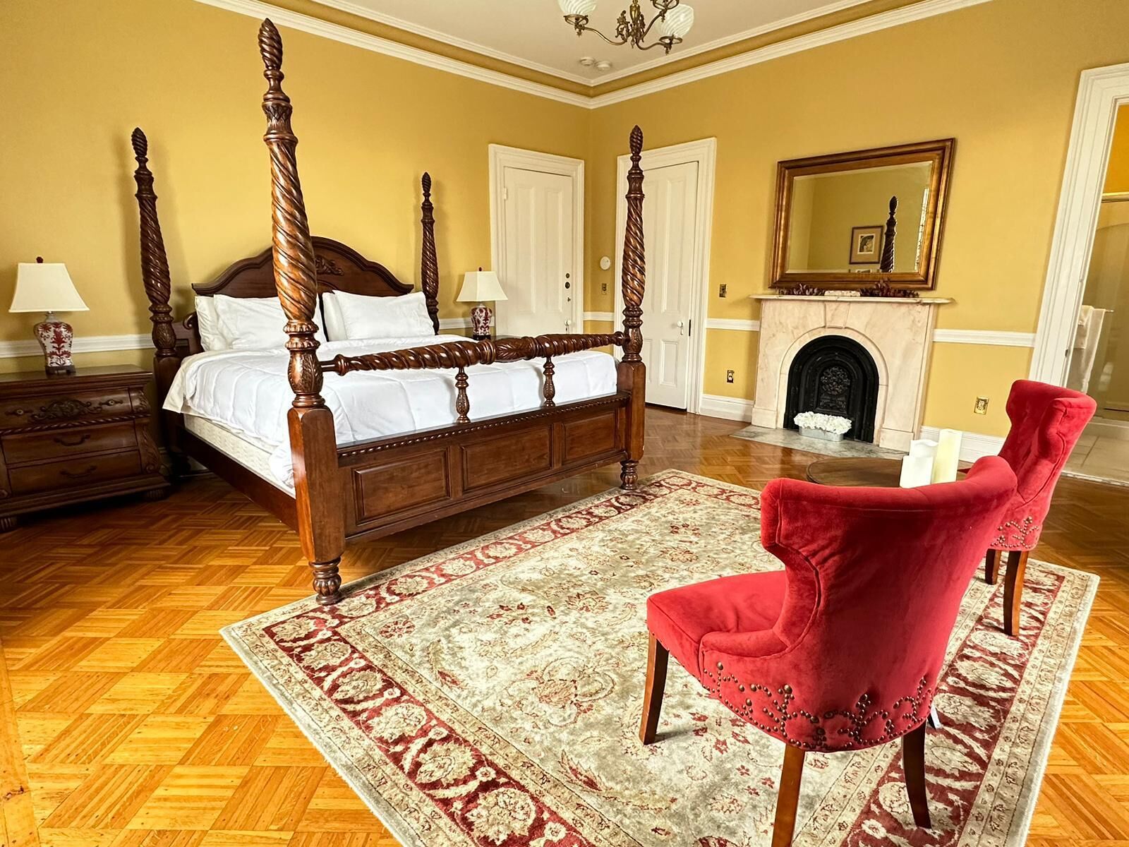 A bedroom with a four-poster bed, a red velvet chair, a fireplace, and a large mirror.