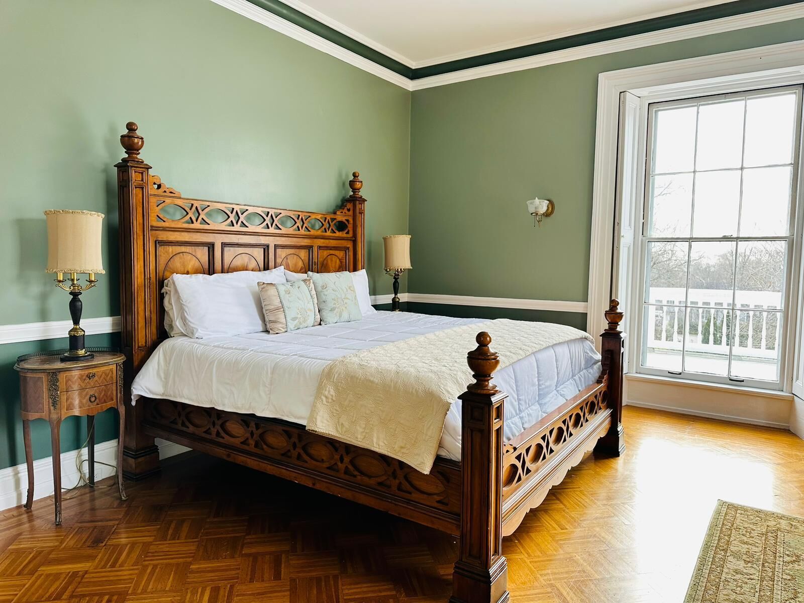 A large bed with a wooden headboard and footboard, flanked by two nightstands with lamps, in a room with green walls and a large window.