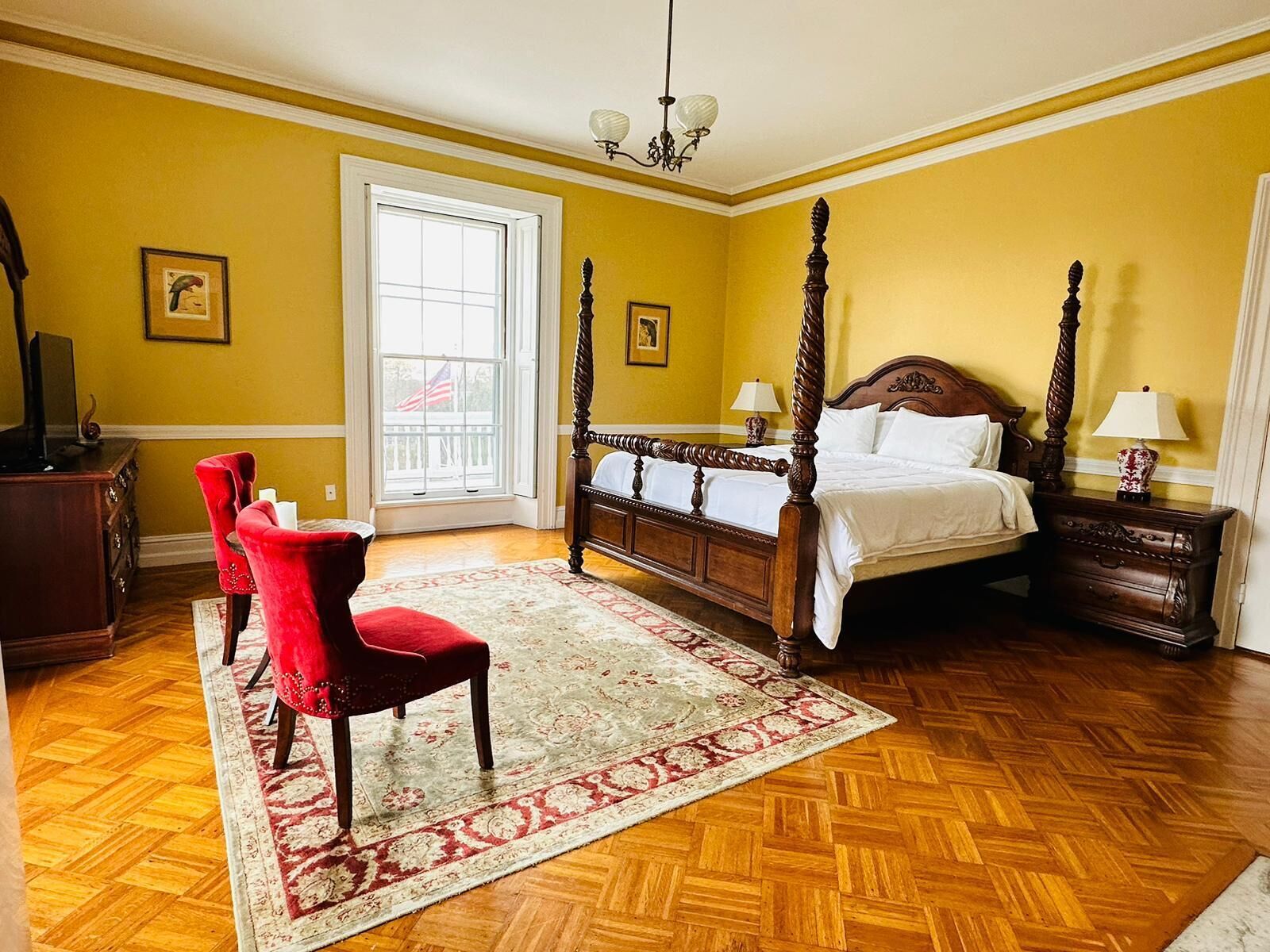 A bedroom with a four-poster bed, a red chair, a wooden dresser, and a large window with an American flag.
