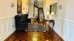 A hallway with a wooden parquet floor, a black upholstered bench, a patterned rug, a chandelier, and a staircase leading to another room. - The Hamilton Hoppin House (Middletown)