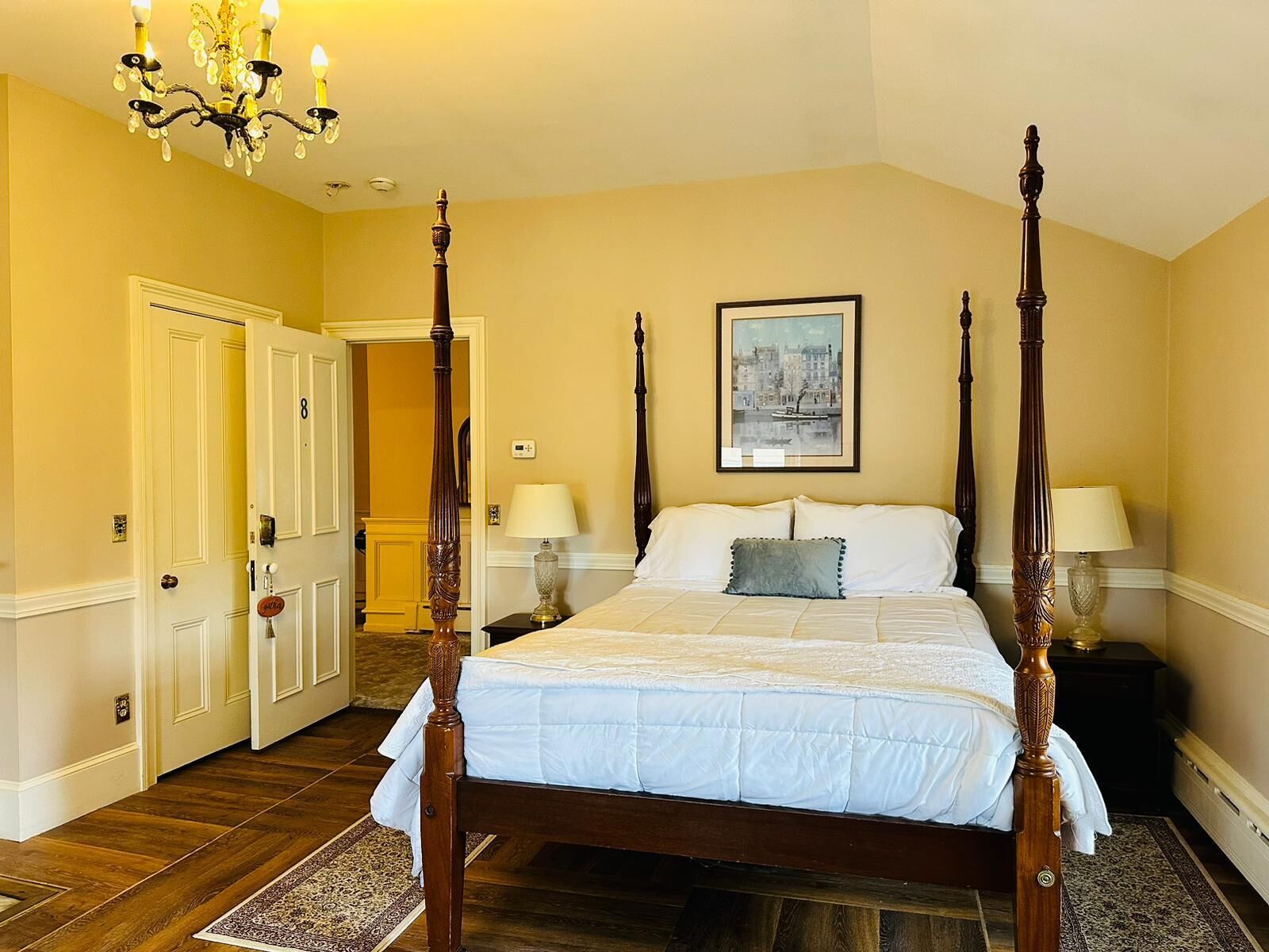 A four-poster bed with a white bedspread and two pillows, a chandelier, and a framed picture on the wall.