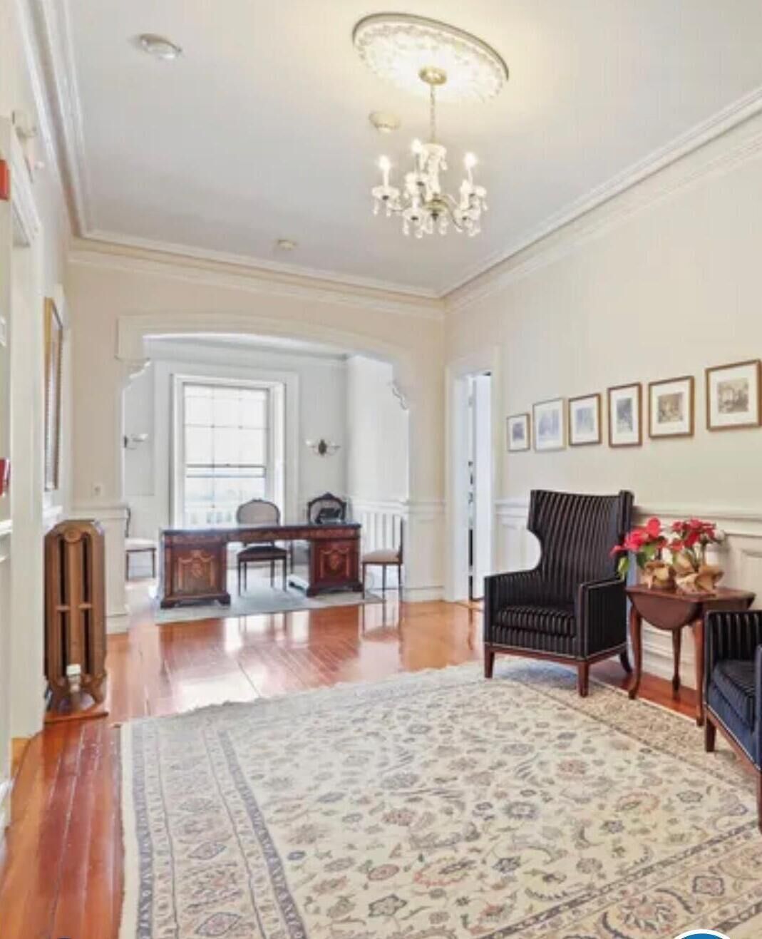 A spacious living room with hardwood floors, a patterned rug, a chandelier, a dark armchair, and a side table with flowers.