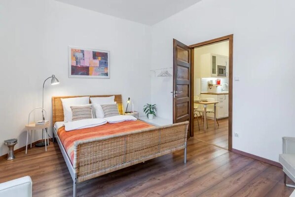 A bedroom with a bed, bedside tables, a lamp, a painting, and a view into the kitchen.