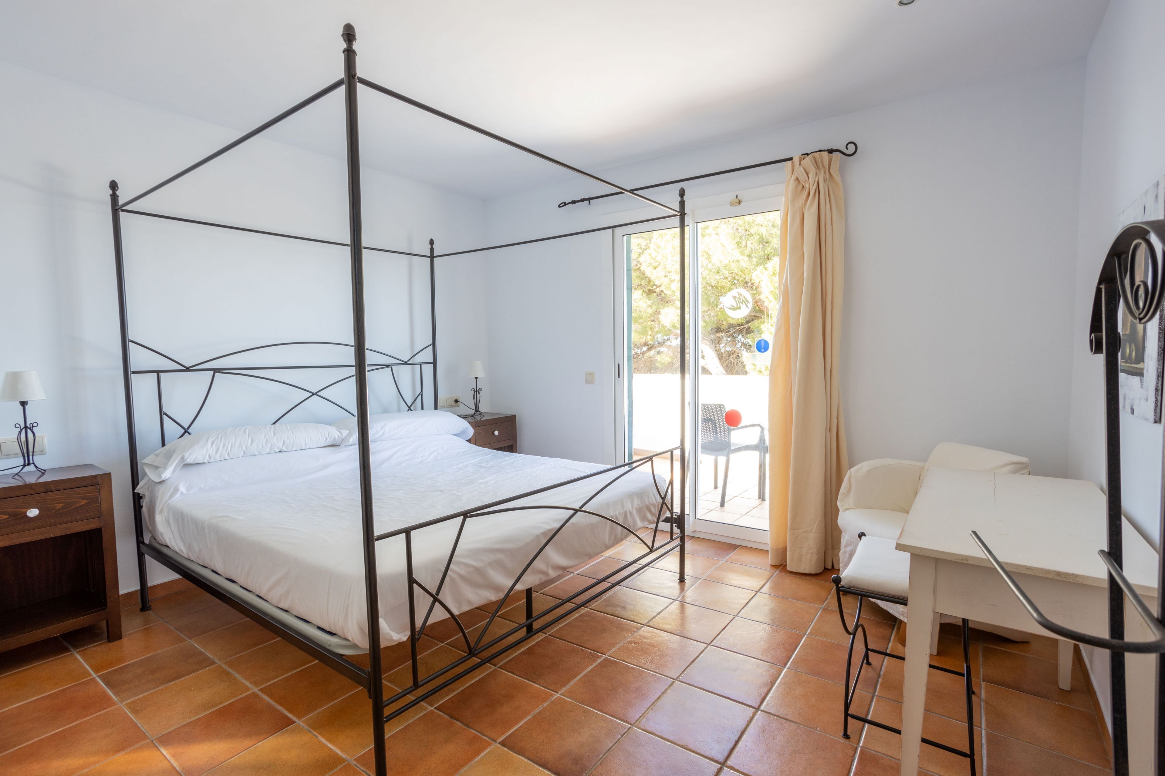 Ein Bett mit Baldachin, mit weißer Matratze und Kissen, ein Nachttisch aus Holz mit einer Lampe, ein weißer Schreibtisch mit Stuhl sowie eine Glastür zum Außenbereich.