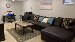 Ein Wohnzimmer mit einem Ecksofa, einem Couchtisch, einem Teppich, einem Schreibtisch mit Computer und einer Wanduhr.