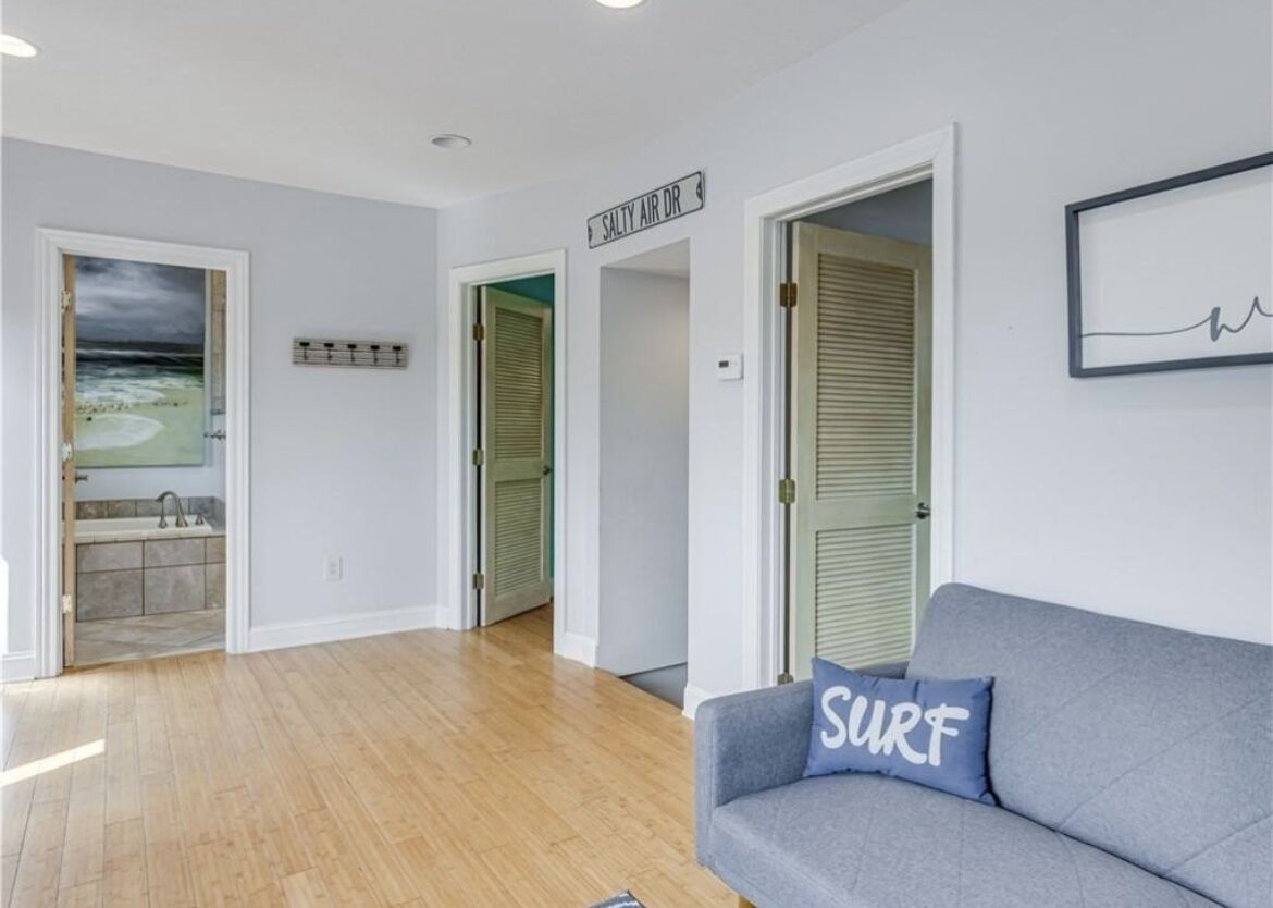 Une pièce avec un plancher en bois, un canapé gris orné d’un coussin sur lequel est écrit « SURF », une porte portant l’inscription « SALTY AIR DR », et un cadre photo fixé au mur.