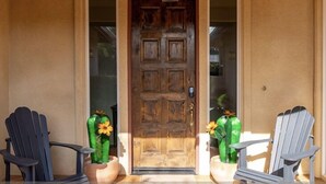 A wooden front door with a glass panel, flanked by two Adirondack chairs with potted plants.