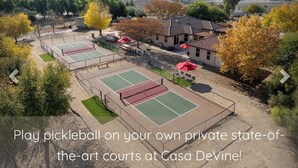A tennis court complex with multiple courts, surrounded by trees and a building with a tiled roof.