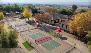 A tennis court complex with multiple courts, surrounded by trees and a building with a tiled roof.