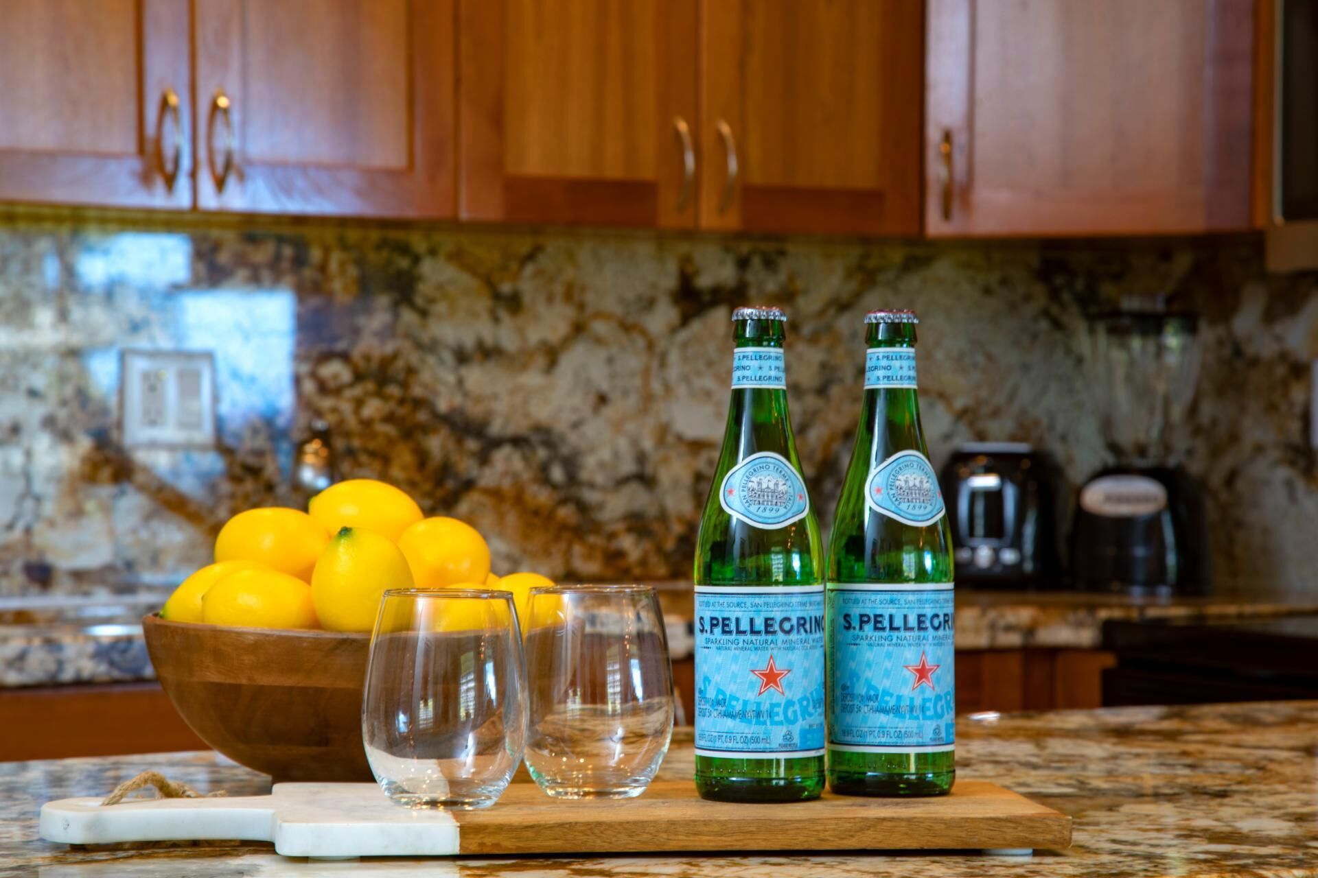 En köksbänk med en träskärbräda, på vilken står två flaskor Pellegrino-kolsyrat vatten, en skål med citroner och två glas.