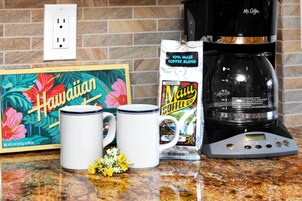 Uma bancada de cozinha com uma cafeteira, duas chávenas, um saco de café e uma caixa com tema havaiano.