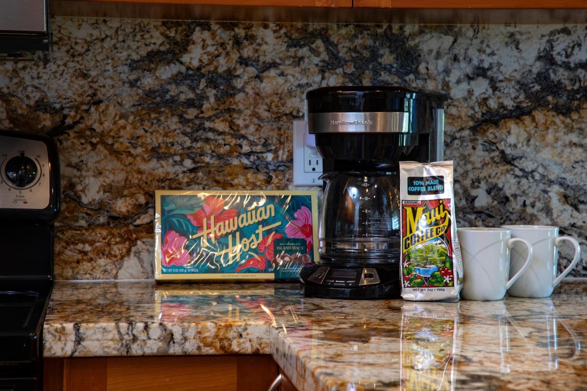 A kitchen counter with a coffee maker, a box of Hawaiian Host coffee, and a mug.