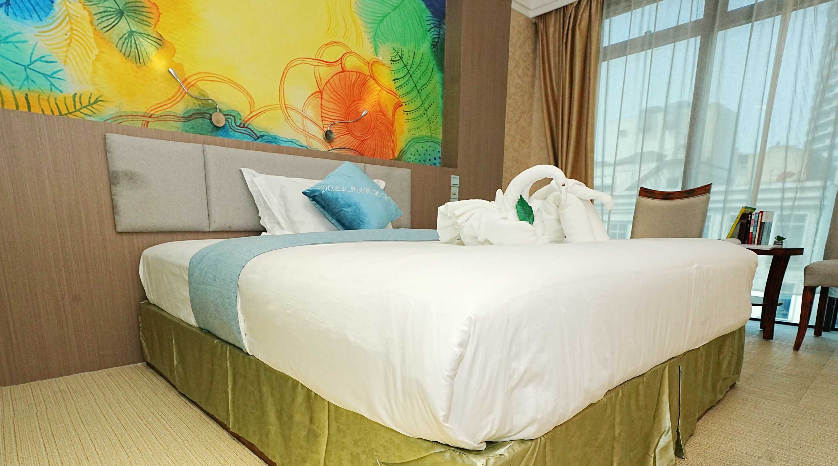 Un dormitorio de hotel con una cama grande, un cuadro abstracto y llamativo en la pared, y vistas de la ciudad a través de amplios ventanales.