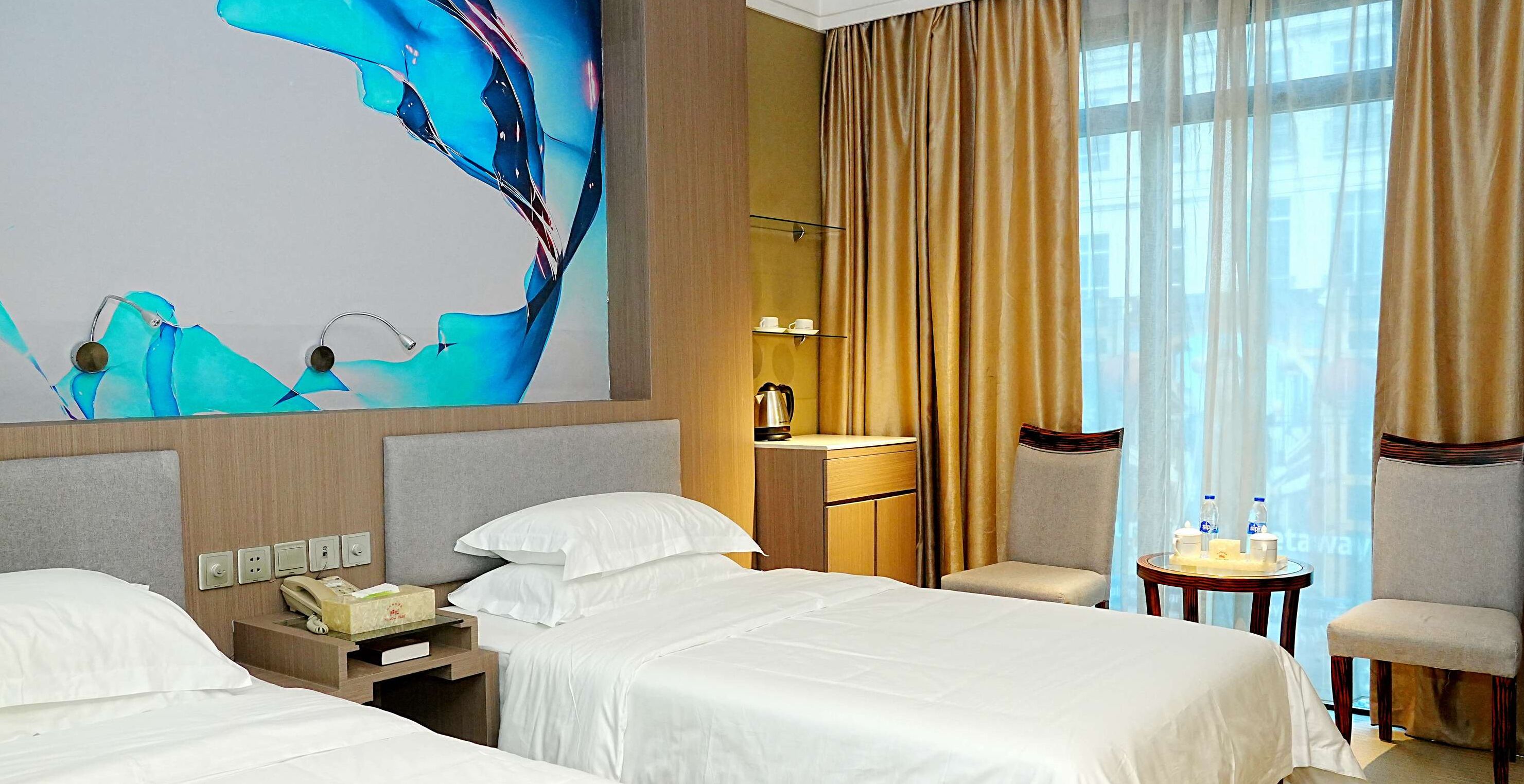 Habitación de hotel con dos camas, un escritorio con teléfono, una silla, una mesita con botellas de agua y un ventanal con cortinas.