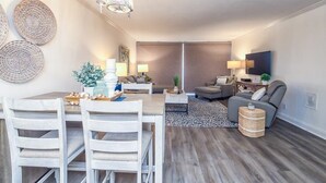 A modern living room with a dining area, featuring a chandelier, sofas, and a TV. - Respite by the Sea The Islander, King Bed, Oceanfront Condo, Pool, Tennis,Cabana (Wrightsville Beach)