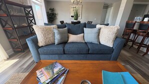 A living room with a dark blue sofa, a coffee table with magazines and a tin, and a 'Welcome' sign on a wooden shelf. - Grand Haven Getaway Welcomes You (Grand Haven)