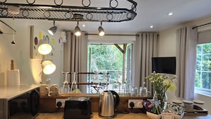 A modern kitchen with a microwave, toaster, kettle, and glassware on the counter. There is a large window with curtains, a ceiling light fixture, and a flat-screen TV mounted on the wall. - Ty Barddu (Newcastle Emlyn)