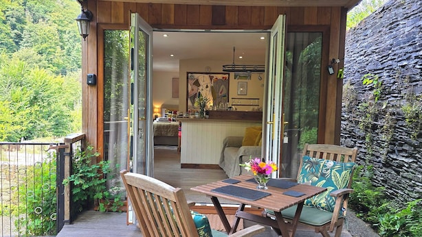 A wooden deck with a table and chairs, a glass door leading to a bedroom, and a stone wall. - Ty Barddu (Newcastle Emlyn)