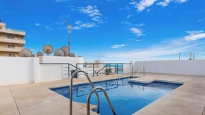 A rooftop swimming pool with a clear blue sky, surrounded by buildings and satellite dishes.