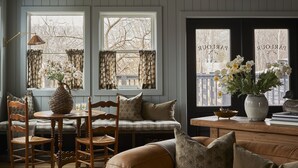 A cozy living room with a brown leather sofa, wooden dining table, and chairs. There are two windows with curtains, a vase with flowers, and a wooden ceiling. - The June Motel, Beaver Valley (Grey Highlands)