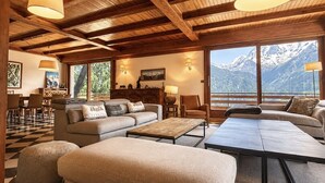 A spacious living area with a large sofa, ottomans, and a coffee table. There are multiple windows offering a view of snow-capped mountains. - LA Villa DES Tacounets (Saint-Gervais-les-Bains)