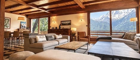 Un vaste espace de vie comprenant un grand canapé, des poufs et une table basse. De nombreuses fenêtres offrent une vue sur les montagnes enneigées.