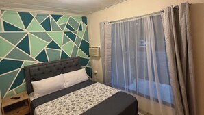 A bedroom with a bed, a nightstand, a geometric-patterned wall, a window with curtains, and a ceiling light. - Oasis by Franklin #2 (La Fortuna)