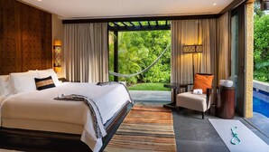 A modern hotel room with a large bed, a sitting area with a chair and table, and a view of lush greenery through large windows.