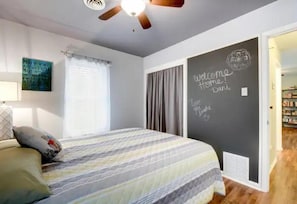 Quarto com cama, ventilador de teto, janela com cortinas e parede com mensagem de boas-vindas.