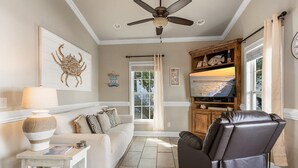 Una sala de estar con sofá, mesa de centro, mueble para televisión con televisor de pantalla plana, ventilador de techo y ventana con cortinas.