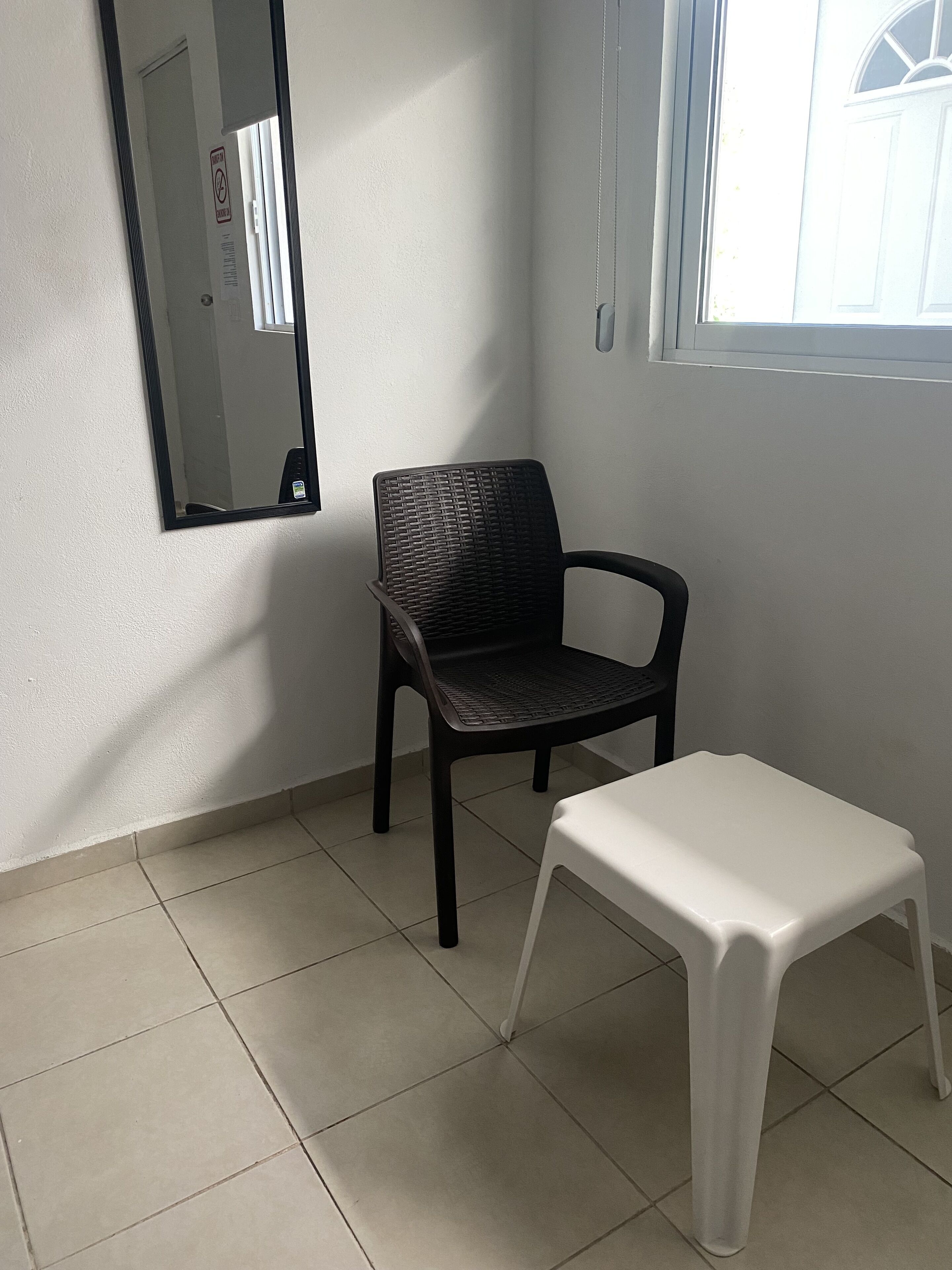 Une pièce avec une chaise noire, un tabouret blanc, un miroir au mur et une fenêtre.