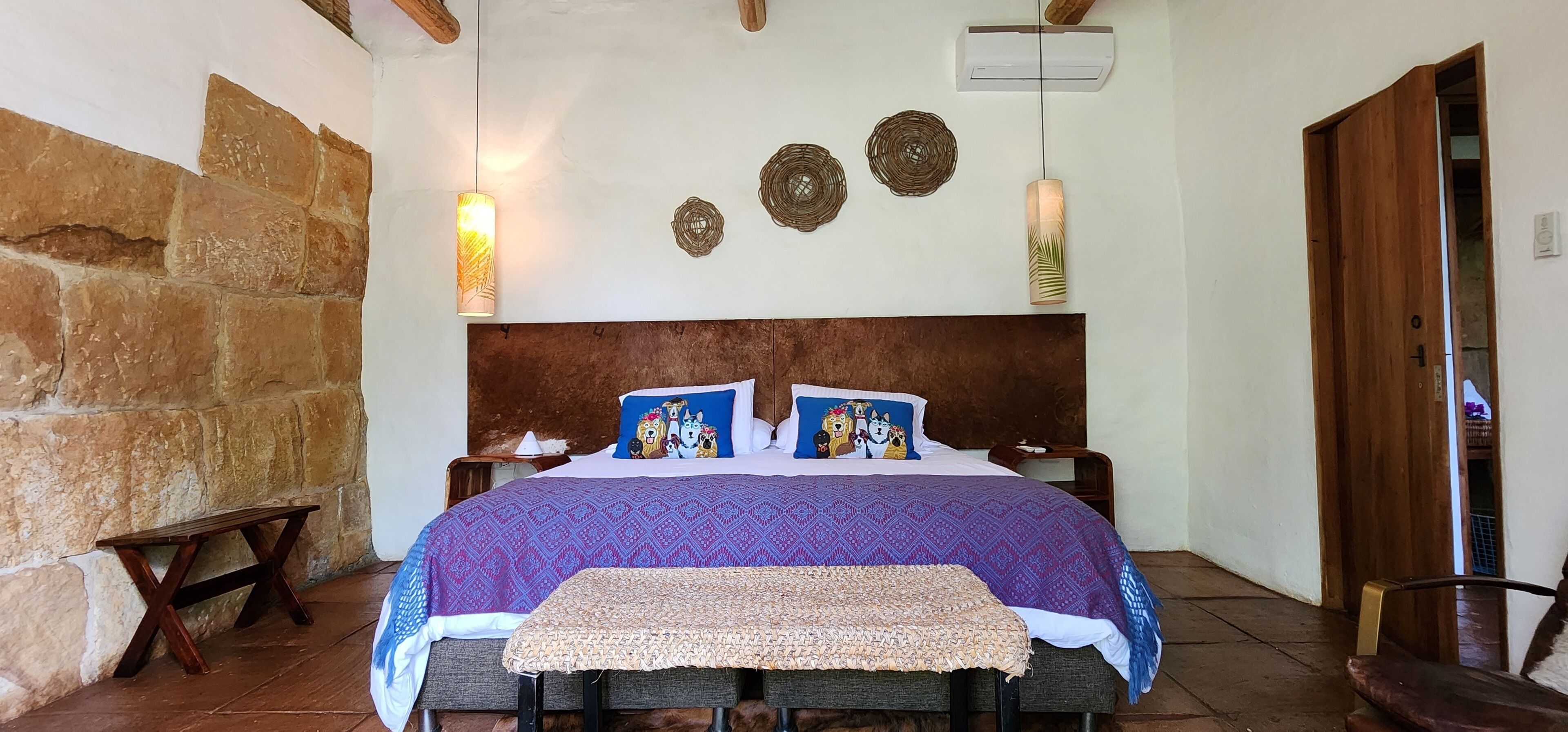 Een slaapkamer met een stenen muur, een houten hoofdbord, een bed met beddengoed met een patroon, twee nachtkastjes en een bankje.