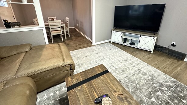 Una sala de estar con un sofá de cuero marrón, una mesa de centro de madera, un televisor de pantalla plana sobre un mueble blanco y una zona de comedor con una mesa y sillas.