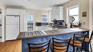 A modern kitchen with white cabinets, a central island with bar stools, a refrigerator, and a microwave. - BEACH PLEASE! Amazing Ocean and Sound Views with Easy Beach Access! Pet Friendly (North Topsail Beach)