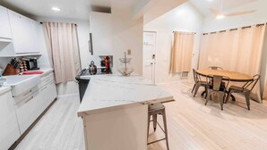Una cocina moderna con armarios blancos, encimera de mármol y comedor con mesa de madera y sillas de metal.