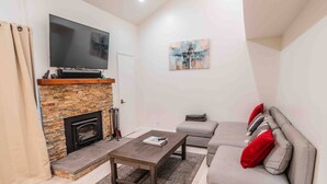 Una sala de estar con chimenea de piedra, un televisor de pantalla plana, un sofá gris con cojines rojos y una mesa de centro.