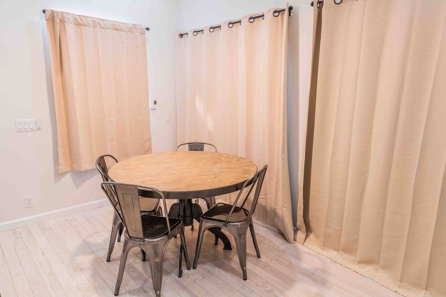 A room with a round wooden table and four chairs, beige curtains, and a light-colored floor.